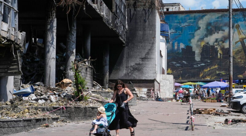 Woman pushes a turquoise stroller with a small child through a rubble-strewn street beside a ruined concrete building and a muraled wall in the background.