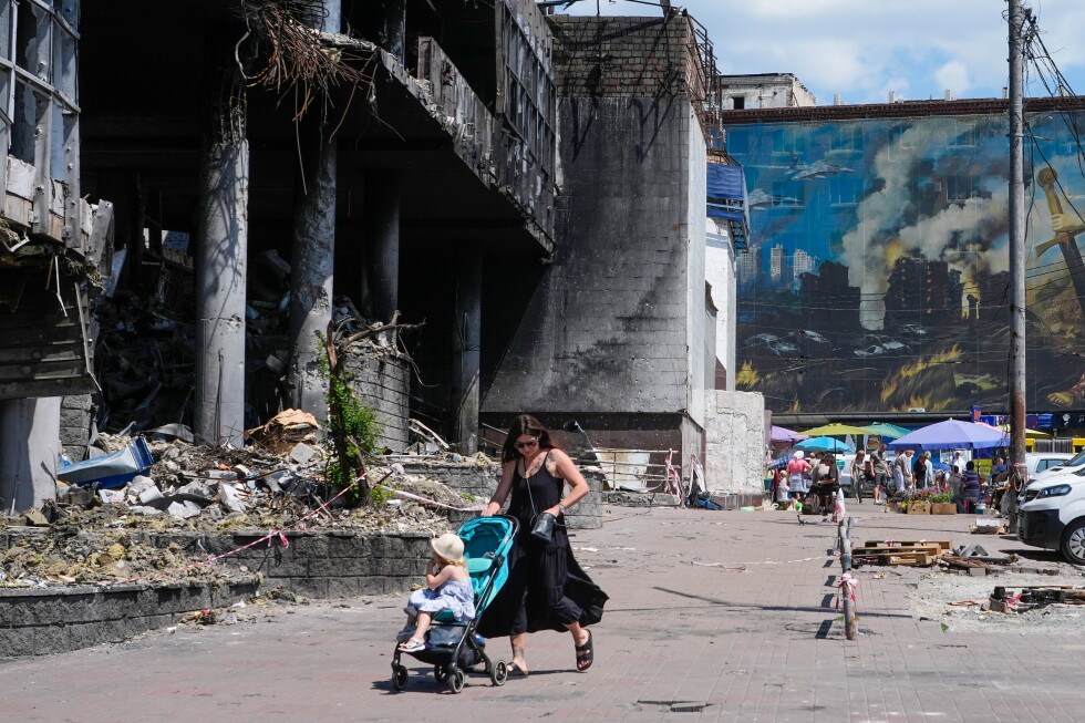 Woman pushes a turquoise stroller with a small child through a rubble-strewn street beside a ruined concrete building and a muraled wall in the background.