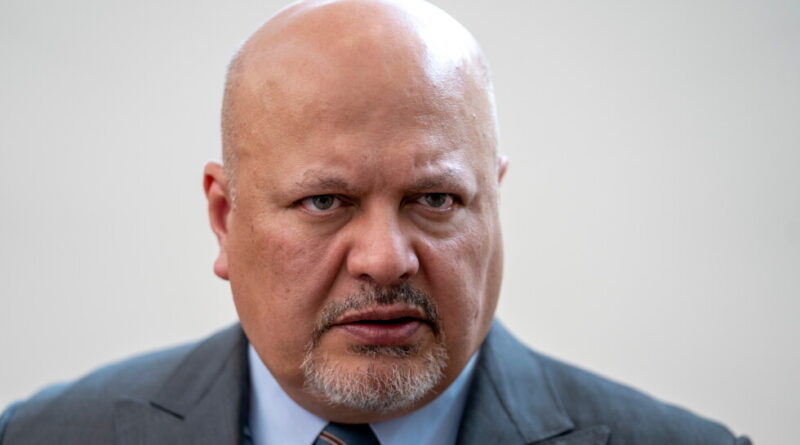 Close-up of a bald man in a gray suit with a blue shirt and striped tie, looking intently at the camera.