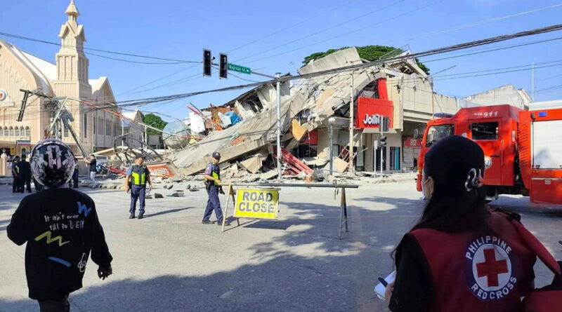 Collapsed building and debris fills a street as responders manage the scene; a red fire truck is on the right, and a 'Road Closed' sign blocks the road.