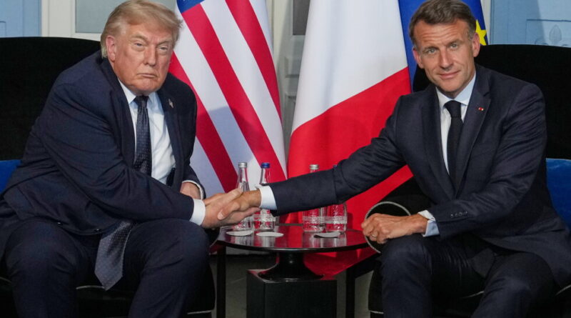 Two male world leaders shake hands across a small table, with US, French, and EU flags in the background.