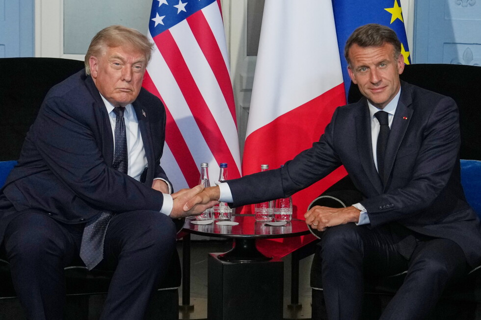 Two male world leaders shake hands across a small table, with US, French, and EU flags in the background.