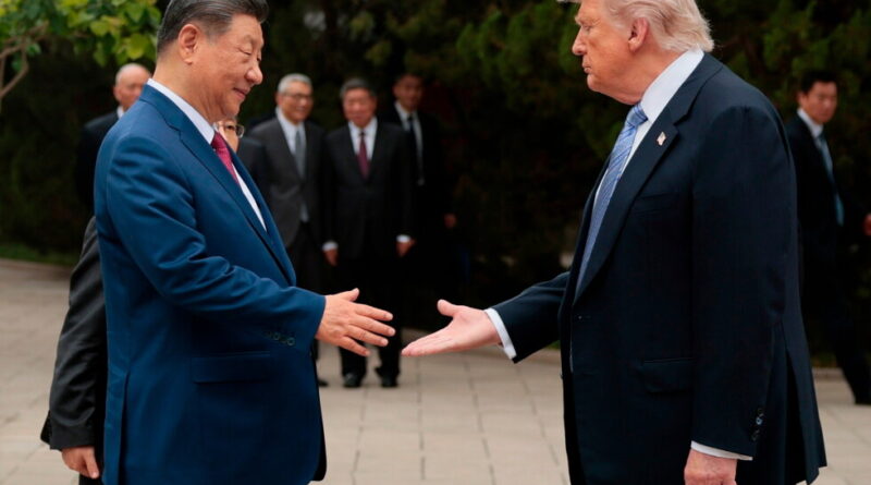 Two men in dark suits shake hands outdoors, with several officials in the background.