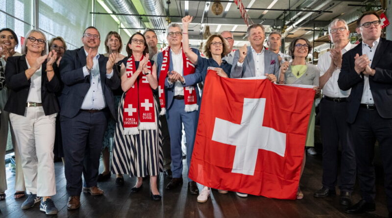 A diverse group of people celebrate indoors, with a large Swiss flag held at center and cheerful expressions.
