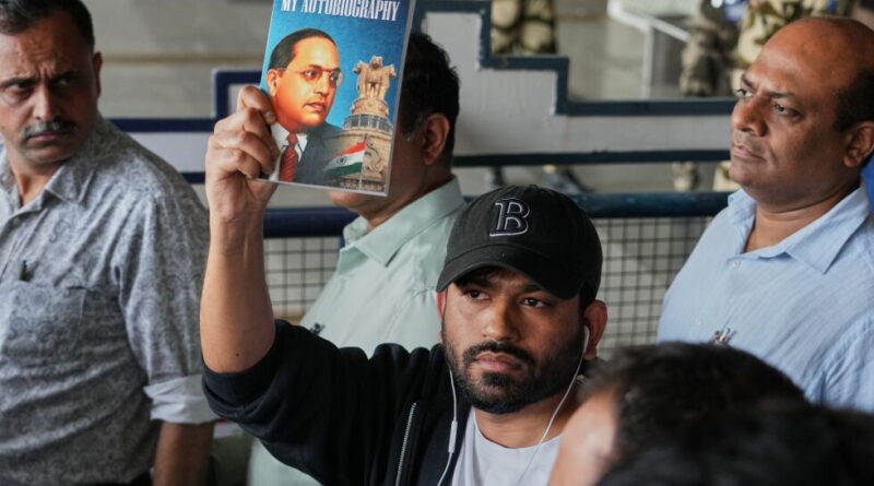 Man in a black cap raises a copy of the book 'My Autobiography' by Dr. B. R. Ambedkar in a crowded setting with onlookers nearby