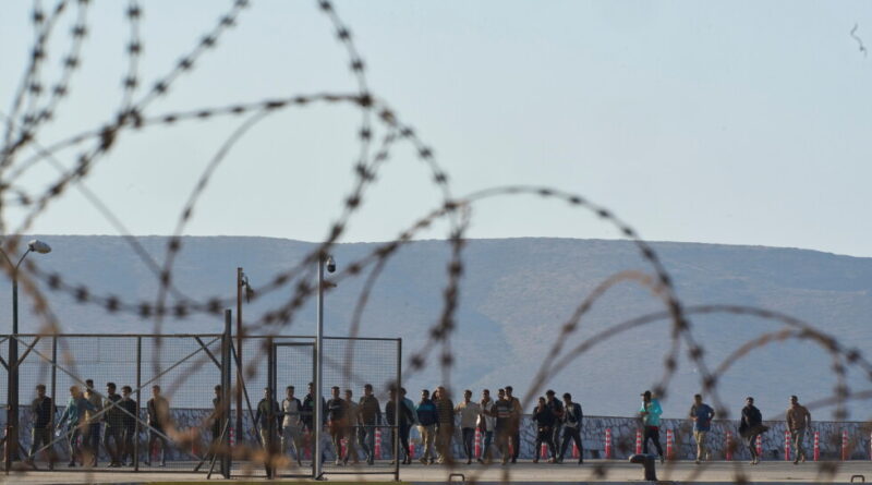 Barbed wire in the foreground; a crowd stands behind a fence at a border area with hills in the distance behind them.