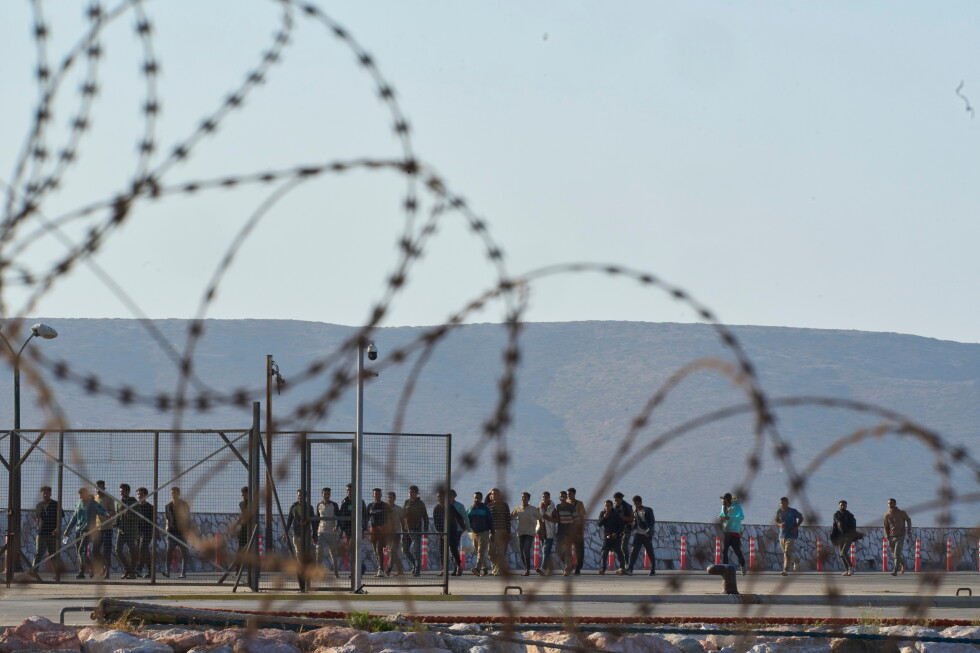 Barbed wire in the foreground; a crowd stands behind a fence at a border area with hills in the distance behind them.