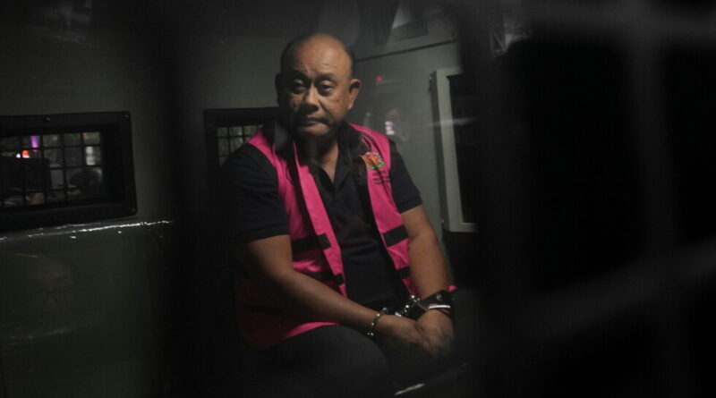 Man in a pink safety vest seated in a dim vehicle, hands cuffed in front, looking toward the camera.