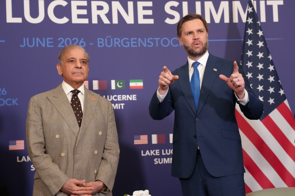 Two men in suits stand on stage at a Lucerne Summit backdrop; one gestures with hands while the other looks ahead, with an American flag nearby.