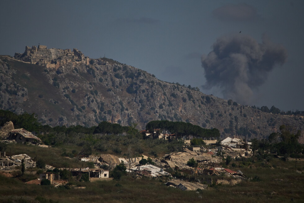 Rugged hillside with an ancient fortress on the left, a dark smoke plume rising on the right, and a small village in the foreground.