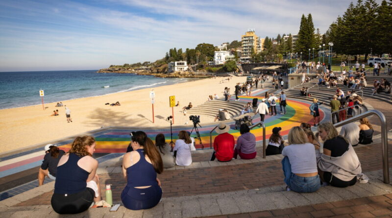 Crowd seated on rainbow-colored circular steps by the beach, watching an outdoor event with cameras nearby and the ocean to the left.