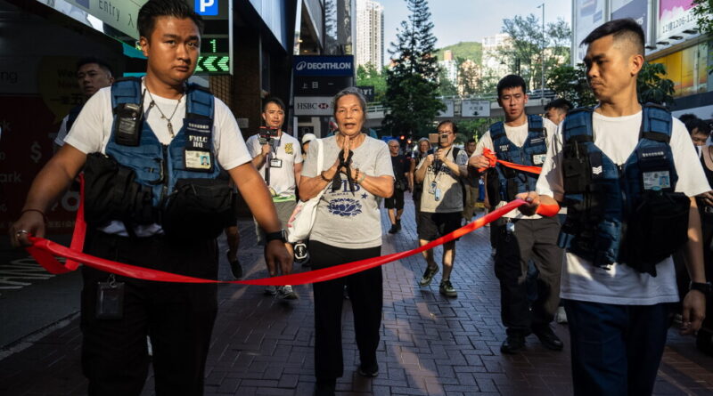 Elderly woman in gray shirt stands with hands pressed together as volunteers hold a red finish-line ribbon on a city street race.