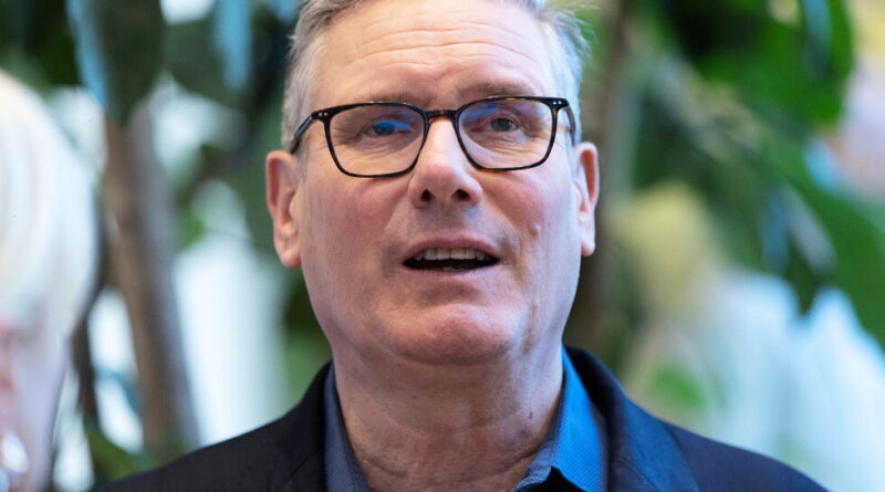 Close-up of a middle-aged man with gray hair and black-rimmed glasses speaking, wearing a dark blazer, with greenery in the background.