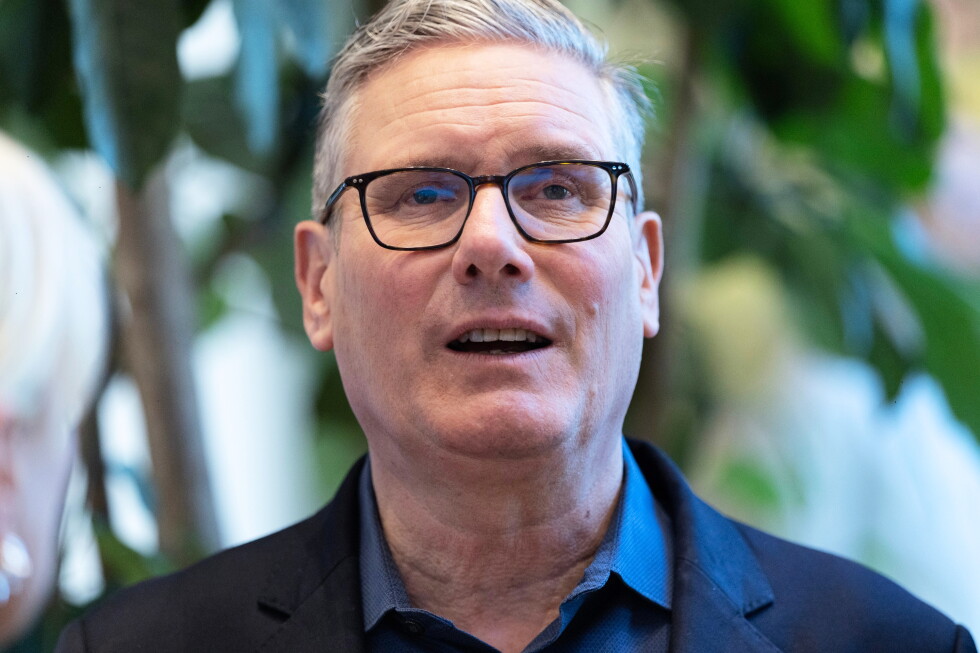 Close-up of a middle-aged man with gray hair and black-rimmed glasses speaking, wearing a dark blazer, with greenery in the background.