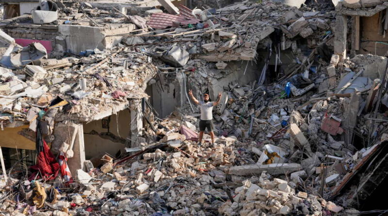 Man stands with arms raised amid a landscape of collapsed buildings and rubble after a disaster, debris scattered everywhere.