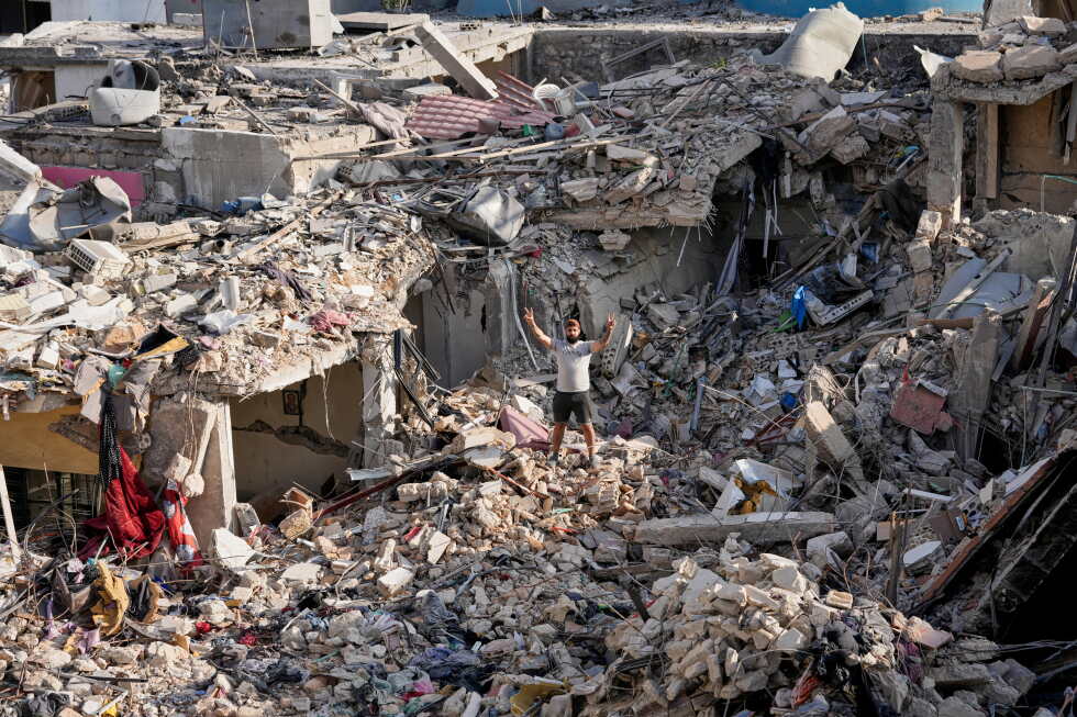 Man stands with arms raised amid a landscape of collapsed buildings and rubble after a disaster, debris scattered everywhere.