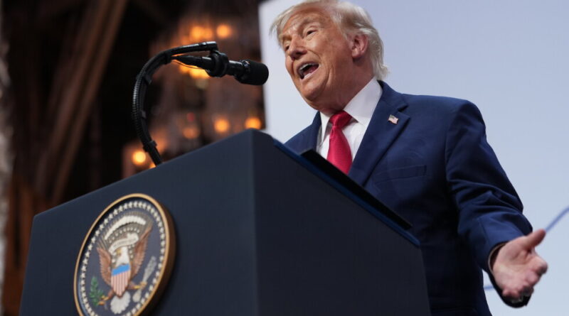 Man in a blue suit and red tie speaks at a podium with a large seal, gesturing with one hand.