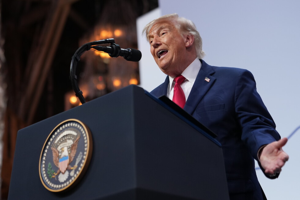Man in a blue suit and red tie speaks at a podium with a large seal, gesturing with one hand.