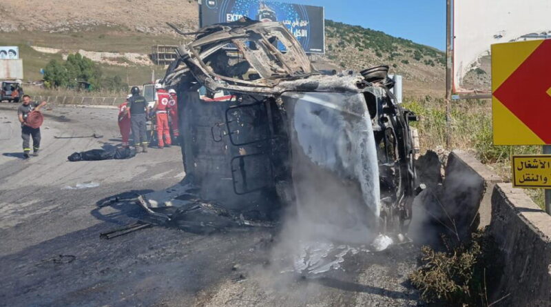 Overturned, severely burned vehicle on a highway with responders in red and black uniforms nearby, smoke rising from the wreckage.