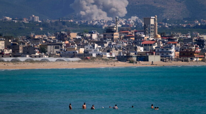 Coastal town along a sandy beach with turquoise water and several people swimming near the shore.