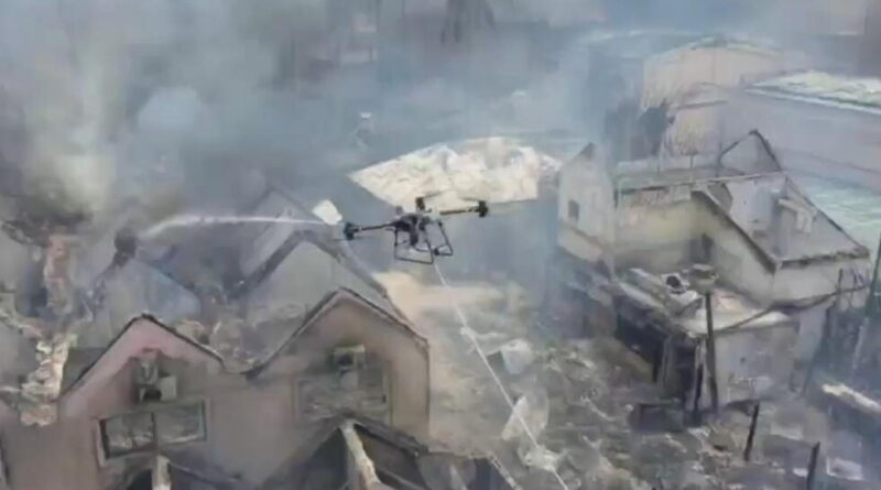 Drone spraying water over a burning village with smoke and damaged houses.