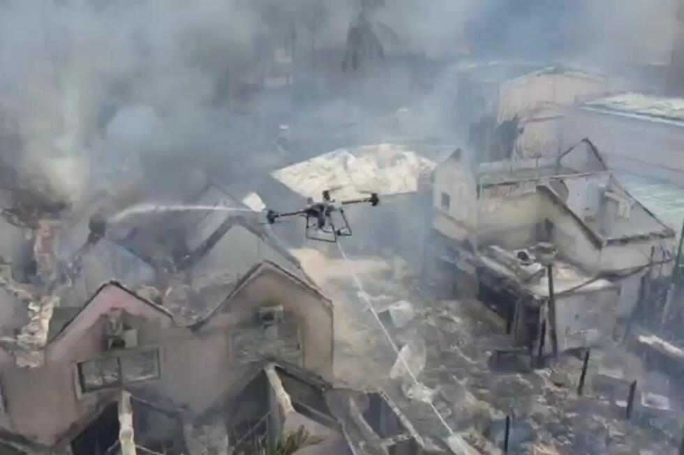 Drone spraying water over a burning village with smoke and damaged houses.