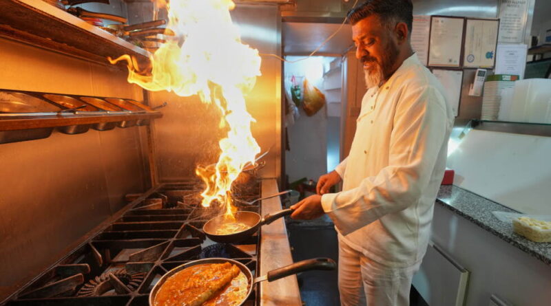 Chef in a white uniform cooks on a gas range, a large flare leaping from a skillet in a busy commercial kitchen.