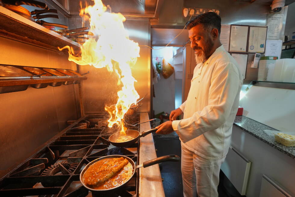 Chef in a white coat flambéing a pan on a commercial stove with bright flames in a busy kitchen.