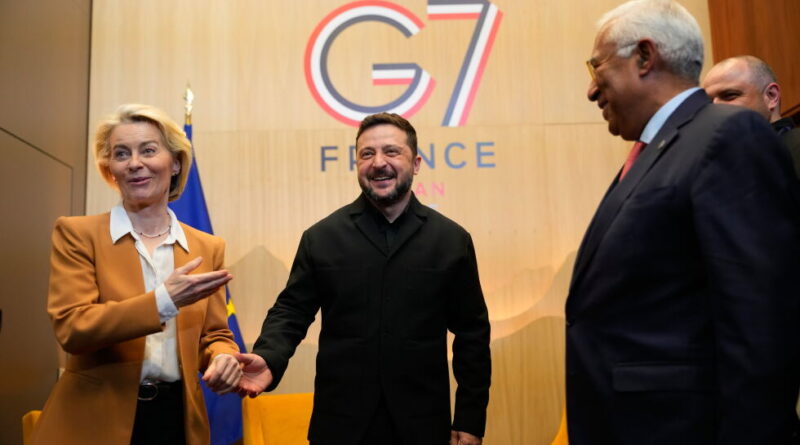 Diplomatic event: a man in black suit shakes hands with a blonde woman in an amber blazer, smiling, as a third man in a suit watches in front of a G7 France backdrop.