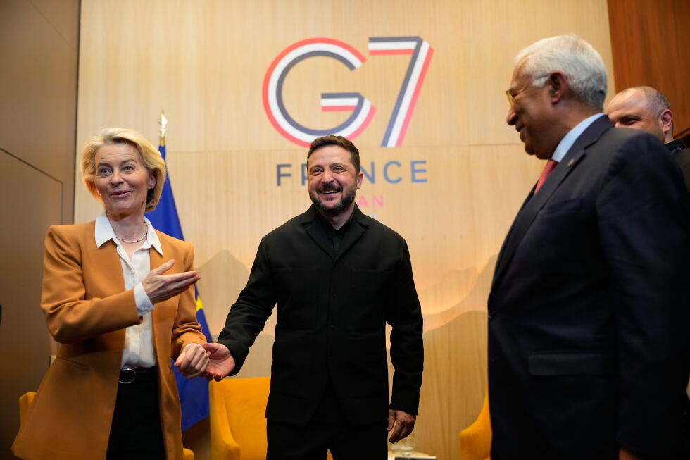 Diplomatic event: a man in black suit shakes hands with a blonde woman in an amber blazer, smiling, as a third man in a suit watches in front of a G7 France backdrop.