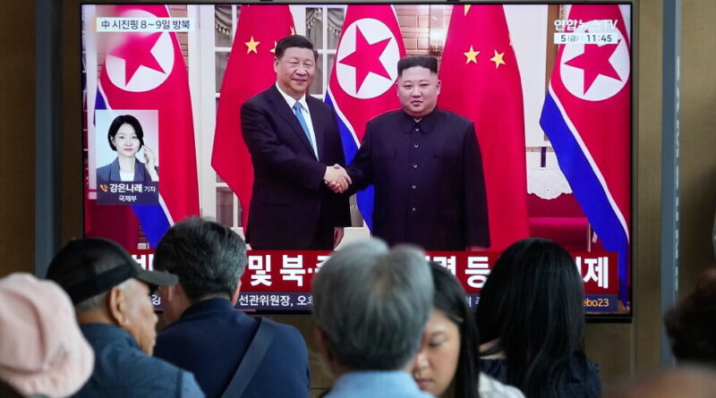Television news screen showing Xi Jinping and Kim Jong-un shaking hands, flanked by Chinese and North Korean flags, with a crowd watching in the foreground.