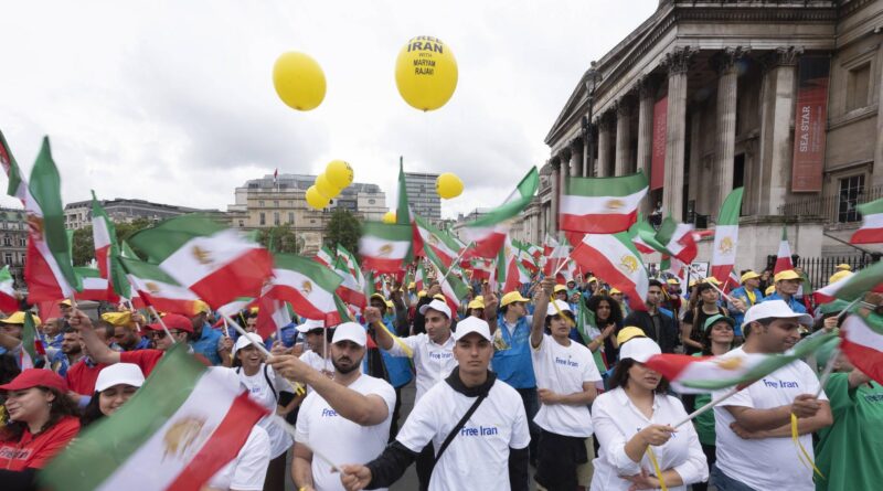 Crowd at a street protest waving Iranian flags with yellow balloons overhead and a historic building in the background.