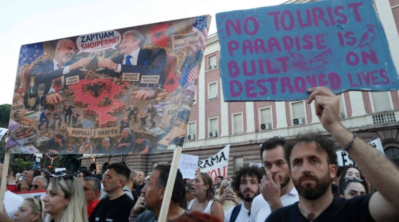 Crowd of protesters holding signs; a blue placard reads, 'NO TOURIST PARADISE IS BUILT ON DESTROYED LIVES'.