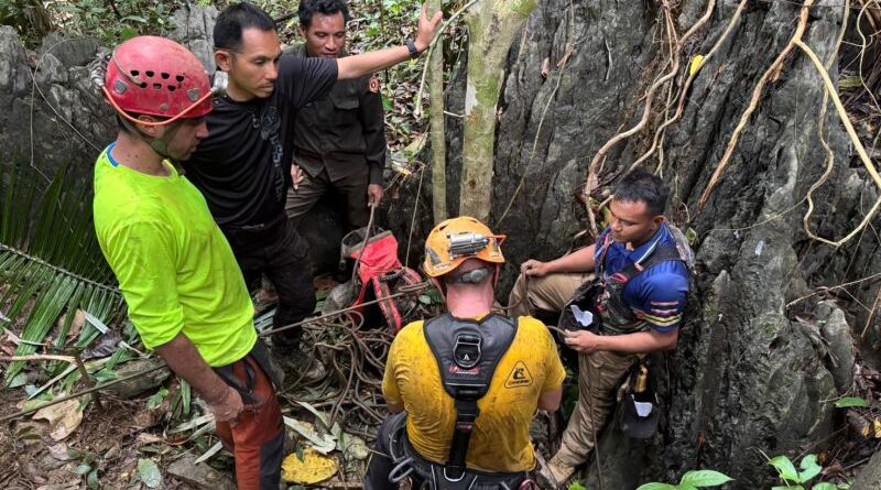 Rescue team in helmets and harnesses gathers in a narrow, vine-covered rocky crevice, coordinating a rope rescue.
