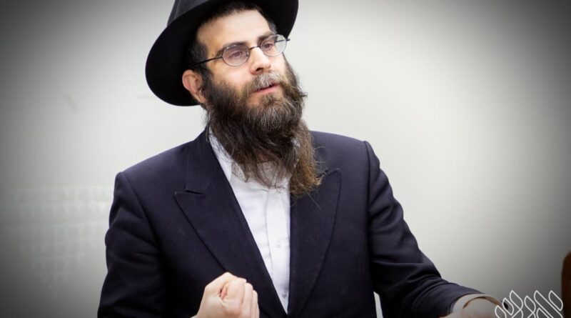 Bearded man in a dark suit and hat speaking at a wooden podium, gesturing with his right hand in a presentation setting